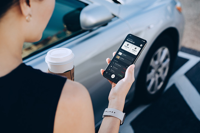 这是一位年轻的亚洲女商人，一边喝着咖啡，一边用智能手机上的移动应用管理着网上银行。转钱，付账单，查看余额，走到她的车在市中心的停车场图片下载